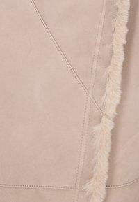 Tissu en suédine beige avec une bordure en fourrure le long des coutures, présentant des coutures visibles et une texture lisse. Le design comprend des panneaux angulaires.