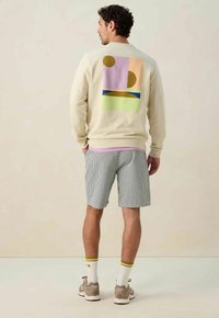Sweatshirt beige avec des graphiques multicolores abstraits au dos, associé à un short rayé. Présente des poignets côtelés et des accents de ourlet rose.