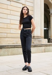 Fekete bordázott cropped póló, magas derekú fekete farmernadrág és fekete platform cipők. A modell a zsebeibe tett kezekkel áll egy kő háttér előtt.