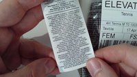 Vitt tvättetikett i tyg med svart text, som beskriver materialkomposition (polyester, elastan) och produktinformation, hålls mellan fingrarna.