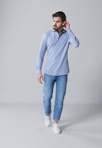 Lichtblauw langlopende polo met een kraag. Draagt het met lichtblauwe spijkerbroek en witte sneakers. Model staat voor een neutrale achtergrond.