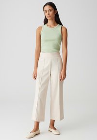 Lichtgroene mouwloze top, crème hoge taille brede broek met voorplooien, en crème instapschoenen tegen een neutrale achtergrond.