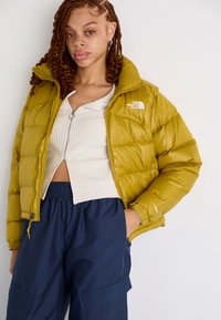 Olivnozeleni pufi jakna z visokim ovratnikom, zadrgo in logotipom na levi prsni strani. Ujemajo se z rebrastim majico in temno modrimi cargo hlačami.