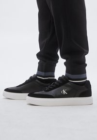 Ténis pretos de couro e camurça, com sola de borracha branca e detalhe de logotipo prateado. Combinados com calças de treino pretas e meias às riscas.