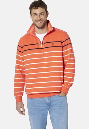 Lächelnder Mann, der einen orangefarbenen Pullover mit Viertelreißverschluss mit weißen und dunklen horizontalen Streifen und hellblauer Jeans trägt, Hand in der Tasche.