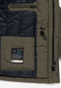 Vista interna di una giacca impermeabile verde con una tasca con zip, un gancio a clip e un'etichetta traspirante impermeabile sulla fodera nera.