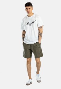 Camiseta blanca de algodón con el texto "Reell", combinada con pantalones cortos verde olivo. El modelo lleva zapatillas blancas y grises, mostrando tatuajes en las piernas.