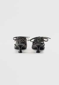 Chaussures à talons noires avec un design à lacets, présentant des clous métalliques à la cheville et un talon effilé. Texture lisse avec une finition brillante.