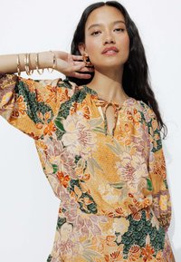 Blouse à motif floral dans des tons chauds d'orange, de vert et de rose, avec une coupe ample, un détail de col à nouer et des poignets élastiques. Accentué de bijoux en or.