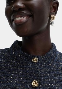 Giacca di tweed blu navy con colletto, decorata con bottoni a nodo dorati e peculiari paillettes. Orecchini dichiarativi in oro e argento completano il look.