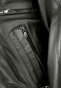 Svart läderjacka med en dragkedjeficka på vänster sida med silverdetaljer, slät textur och strukturerad design.