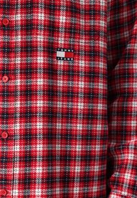 Czerwona i czarna koszula flanelowa w kratę z białymi akcentami, z czerwonymi guzikami i naszywką z logo „TOMMY JEANS” na piersi.