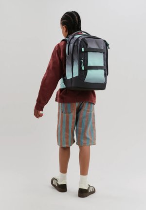 Kind mit geflochtenem Haar, trägt gestreifte Shorts, ein langärmliges burgunderrotes Shirt und schwarze Sneakers, trägt einen großen türkis-schwarzen Rucksack.