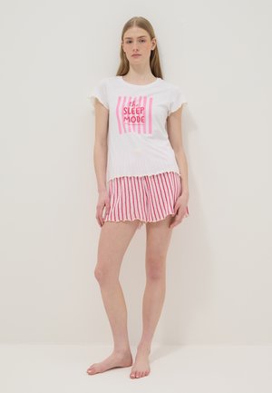 Junge Frau steht barfuß, trägt ein weißes Kurzarmshirt mit pinkem Schriftzug „the sleep mode“ und rot-weiß gestreifte Shorts, neutraler Hintergrund.