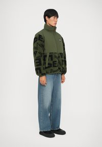 Giacca in pile verde con maniche nere a fantasia, collo alto, chiusura con zip e patch con logo. Abbbinata a jeans larghi blu e scarpe nere.