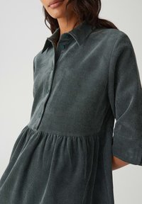 Vrouw in een donkergroene corduroy jurk met knopen aan de voorkant en een geplooide taille, gefotografeerd van midden borst tot heupen.