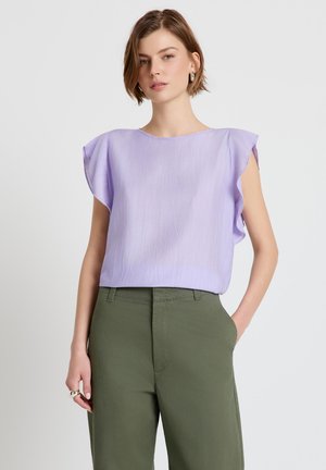 Frau mit kurzen Haaren, die eine hellviolette ärmellose Bluse mit Rüschenschultern und olivgrüne hoch taillierte Hosen trägt, die Hand in der Tasche.