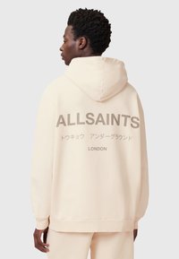 Felpa crema con un grande logo sul retro, con la scritta "ALLSAINTS" e "LONDON". Tessuto morbido, vestibilità comoda e cappuccio con coulisse.