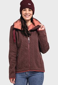 Maronfarbiger Jacke mit Muster und rosa Innenfutter, Frontreißverschluss und verstellbarem Kapuze. Das Modell trägt eine passende Mütze und ein helles Shirt darunter.