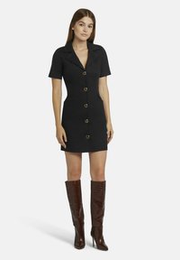 Schwarzes kurzärmeliges Hemdkleid mit figurbetonter Passform, zwei aufgesetzten Taschen vorne und goldfarbenen Knöpfen, kombiniert mit kniehohen braunen Stiefeln im Krokodil-Look.