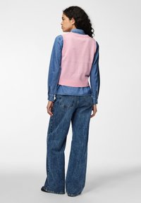Chaleco de punto rosa sobre una camisa de mezclilla, combinado con jeans de pierna ancha azul. Presenta texturas contrastantes y un corte relajado.