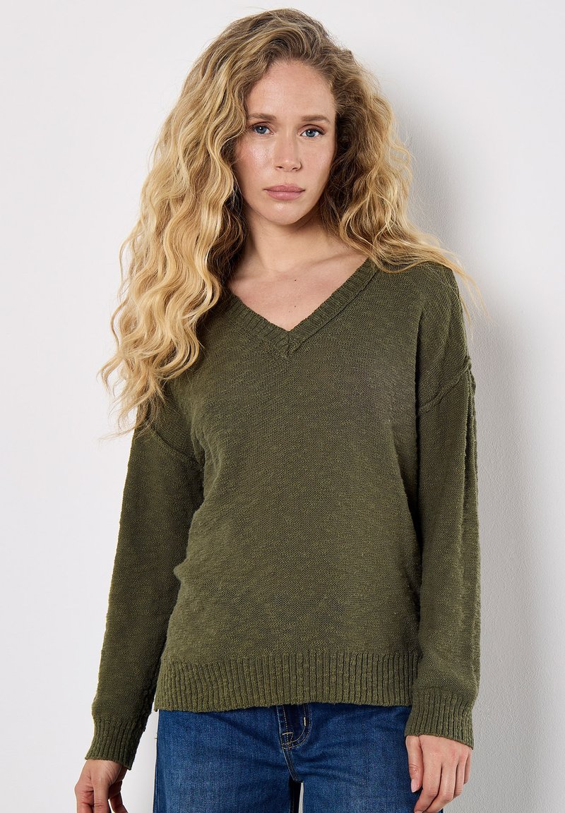 Pull à col en V de couleur vert olive, en tissu tricoté texturé avec des manches longues et des poignets et un ourlet côtelés, associé à un jean bleu.