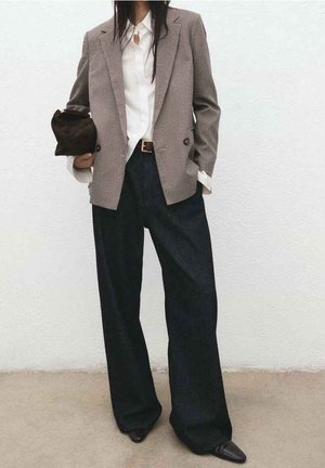 Personne portant un blazer à carreaux, une chemise blanche, un pantalon noir ample, des chaussures pointues, tenant une pochette noire devant un mur uni.