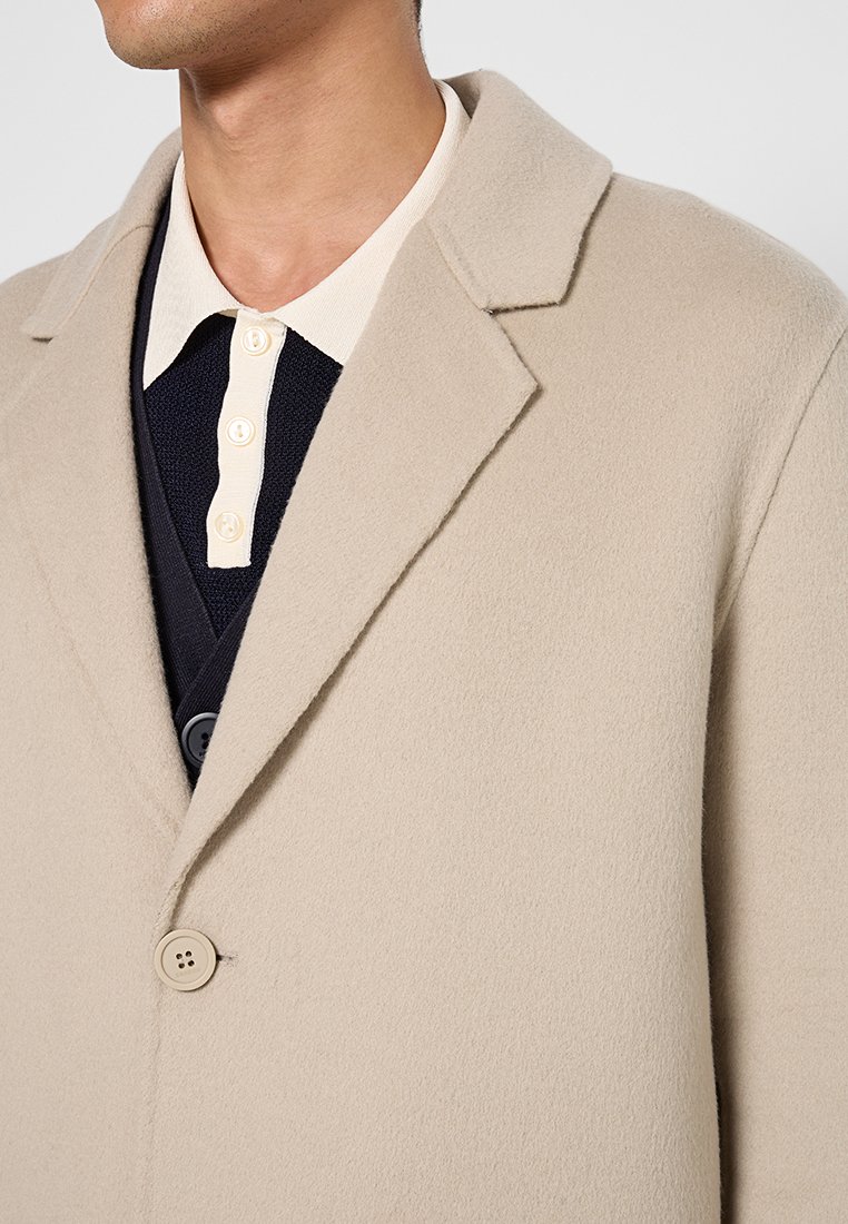 Beige Wollmantel mit Revers, einem einzelnen Knopfverschluss und weicher Textur, über einem marineblauen Pullover mit einem cremefarbenen Kragen getragen.
