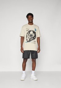 Vyras, dėvintis smėlio spalvos LA Dodgers grafišką marškinėlį, juodas tinklines šortus, baltas kojines ir baltus sportinius batus, stovi prieš neutralų foną.