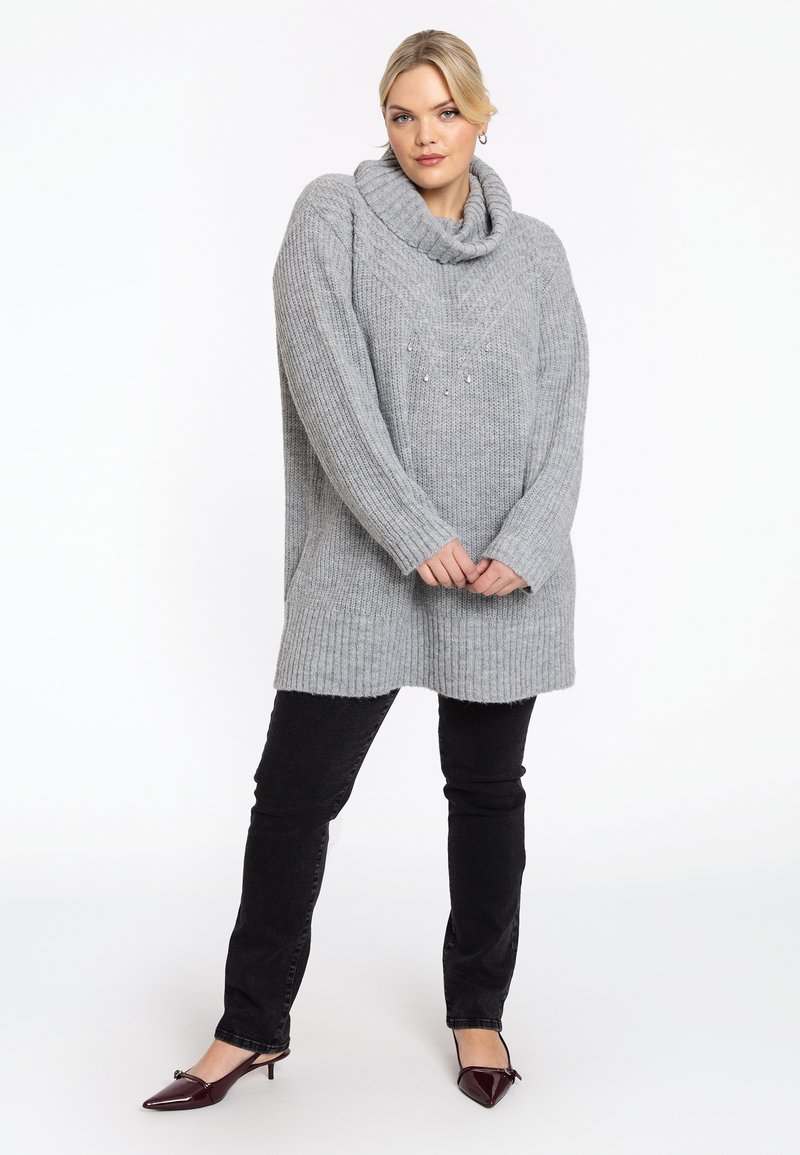 Frau mit blonden Haaren, die einen grauen oversized Strickpullover mit Rollkragen, schwarze Jeans und bordeauxfarbene spitze, flache Schuhe trägt und steht.