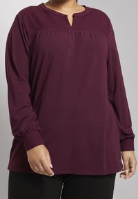 Marmorød langærmet top med smocked yoke og placket-udskæring. Har samlede ærmer og teksturerede manchetter. Blødt, draperet stof.