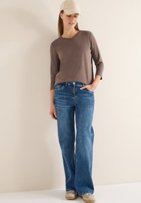 Haut marron à manches trois-quarts associé à un jean bleu à jambes larges. Le mannequin porte une casquette beige et des chaussures claires, debout devant un fond neutre.