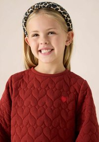 Rode gewatteerde sweatshirt met hartpatroon, voorzien van een kleine rode hartapplicatie. Het kind draagt een haarband met luipaardprint.