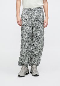 Személy laza, fekete-fehér pöttyös nadrágban és szürke Nike tornacipőben, egyszínű fehér háttér előtt állva.