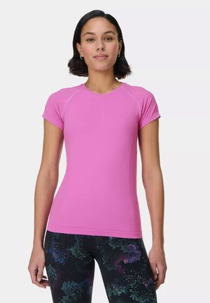 Woman with short dark hair wearing a fitted bright pink short-sleeve shirt and dark patterned leggings, standing against a plain background.