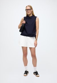 Tumesinist värvi varrukateta pluus kraega, valgete lühikeste pükstega. Mustad tossud kreemikate detailidega ja valged sokid. Lihtne, kaasaegne disain.