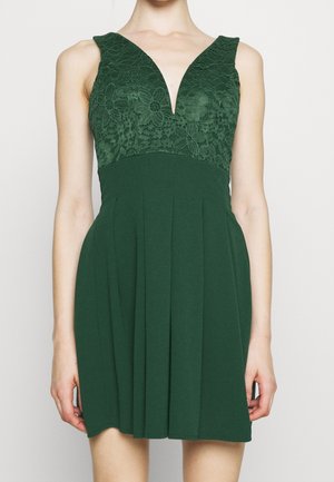 Ärmelloses dunkelgrünes Kleid mit floraler Spitzen-Büste und tiefem V-Ausschnitt, fließendem plissiertem Rock, getragen von einer stehenden Person.