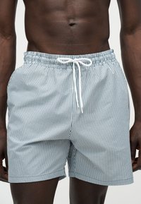 Homme portant un short de bain rayé blanc et bleu marine avec un cordon blanc, montré de la taille jusqu'à la mi-cuisse sur un fond uni.
