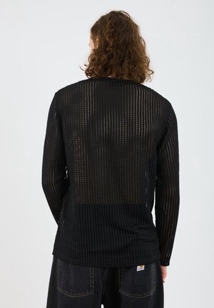 Personne aux cheveux bouclés portant un haut noir à manches longues, semi-transparent avec un motif en grille et un pantalon foncé, vue de dos sur un fond uni.