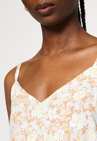 Haut sans manches à motif floral dans un doux ton pêche, doté d'un décolleté en V, de fines bretelles délicates et d'un tissu léger avec une finition légèrement texturée.