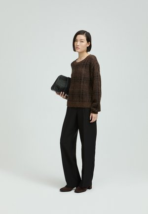 Woman wearing a brown patterned sweater, black wide-leg trousers, and brown shoes, holding a black woven handbag, standing against a neutral background.