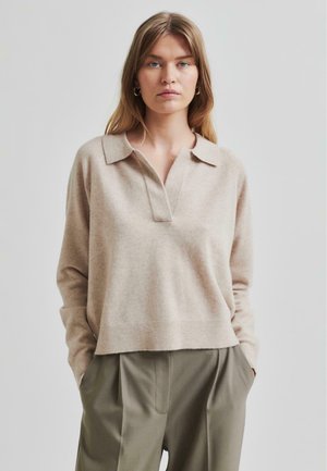 Maglione beige a maglia con colletto, caratterizzato da un'apertura con patta, vestibilità rilassata e orlo a coste. Indossato sopra pantaloni plissettati verde oliva.