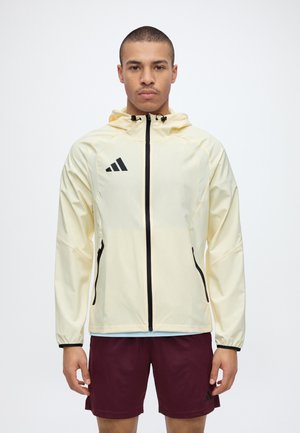 Un homme porte une veste Adidas jaune pâle à capuche et à fermeture éclair, avec un short bordeaux, sur un fond blanc uni.