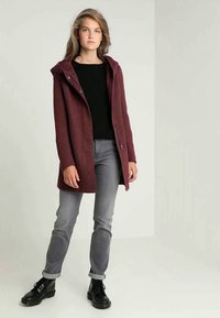 Maroon hooded coat with textured fabric, front buttons, and side pockets. Paired with black top, gray jeans, and black lace-up boots.