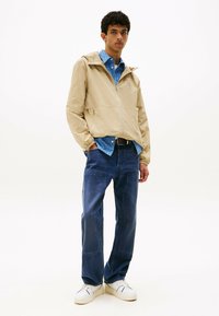 Beżowa, lekka kurtka z kapturem, noszona na niebieską, w kratę koszulę. Niebieskie dżinsy i białe trampki dopełniają stylizację.