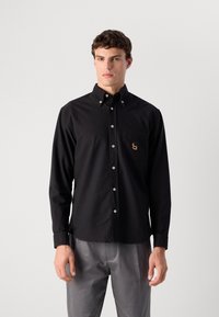 Modelo masculino vistiendo una camisa negra de botones con un logo en el bolsillo del pecho y pantalones grises, de pie contra un fondo claro y liso.