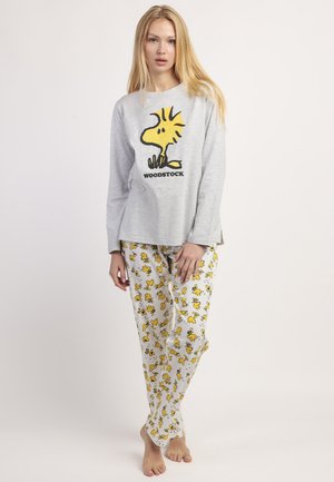 Joven mujer con camiseta de manga larga gris y pantalones de pijama blancos con patrones amarillos del pájaro Woodstock, de pie descalza contra un fondo liso.