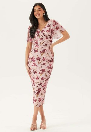 Kvinna som ler, klädd i en rosa blomstrig midiklänning med volangärmar och nudefärgade remsandaler, stående med ena handen på höften.