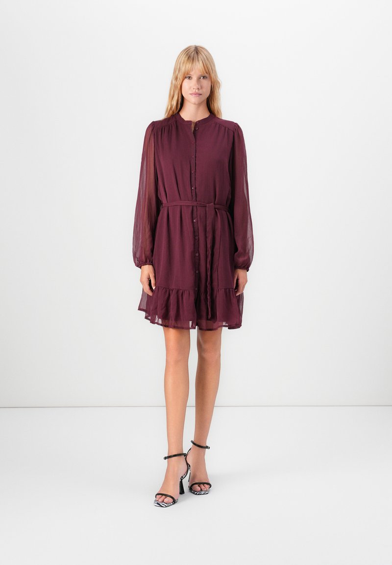 Woman wearing a knee-length burgundy buttoned dress with long sheer sleeves, a waist tie, and black ankle-strap heels with zebra print.
