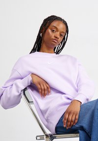 Sweatshirt roxo com decote redondo e punhos canelados, apresentando um pequeno logótipo branco da Nike. O modelo está sentado numa cadeira de metal, vestindo calças de ganga azuis.